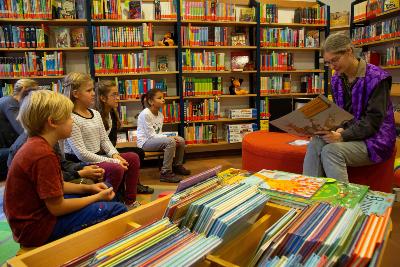 Frau liest mehreren Kindern im Büchereiraum aus einem Bilderbuch vor, Bücherregale im Hintergrund