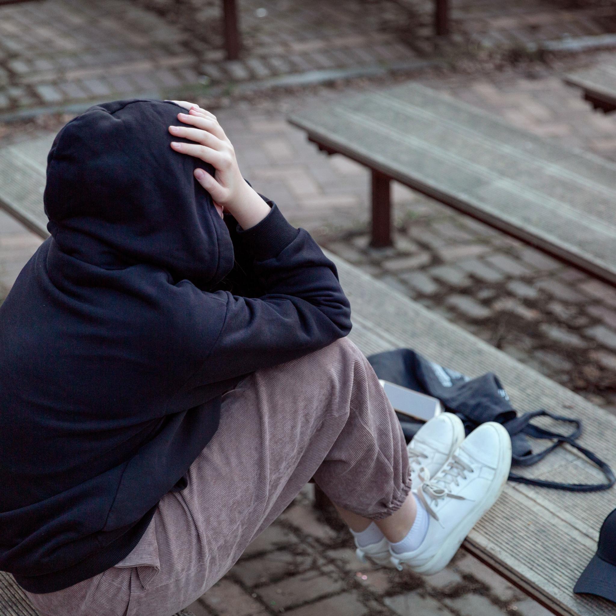 Eine Teenagerin mit schwarzem Hoodie und aufgezogener Kapuze sitzt auf einer Bank. Sie verbirgt ihr Gesicht in ihren Händen. Lebenssituation von Teenagern, die sich einsam fühlen.
