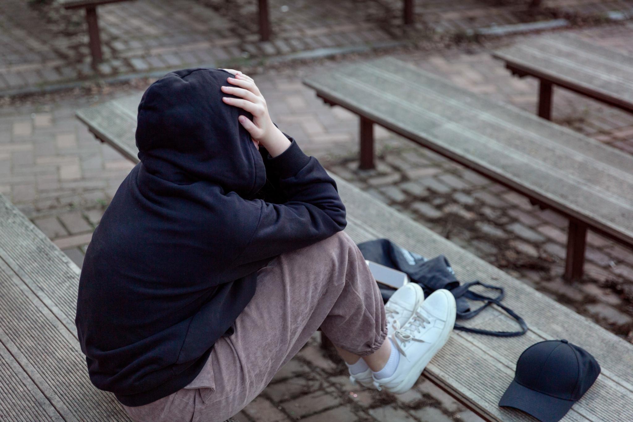 Eine Teenagerin mit schwarzem Hoodie und aufgezogener Kapuze sitzt auf einer Bank. Sie verbirgt ihr Gesicht in ihren Händen. Lebenssituation von Teenagern, die sich einsam fühlen.