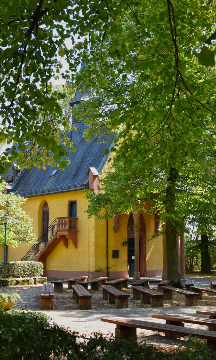 Die gelbe Kapelle versteckt hinter Laubbäumen im Sommer mit vielen Holzbänken davor harmonisch angeordnet