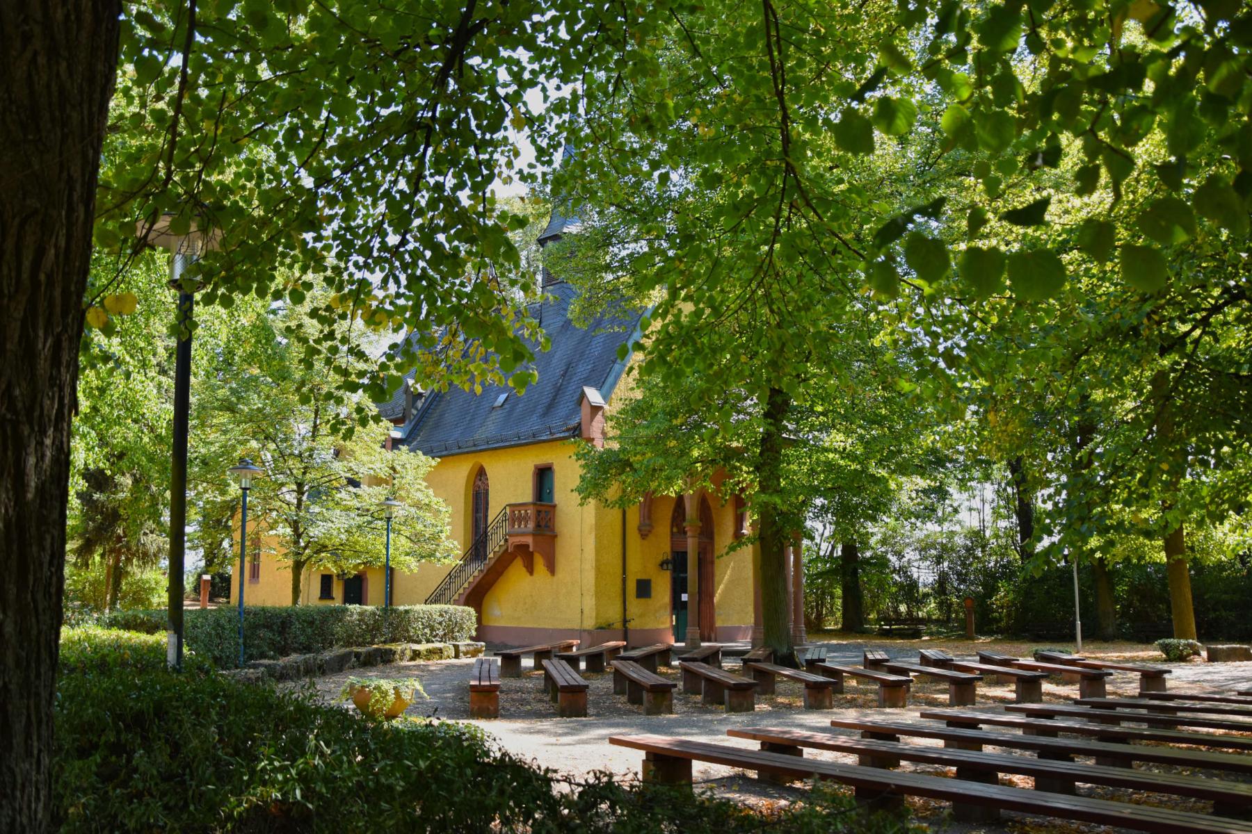 Die gelbe Kapelle versteckt hinter Laubbäumen im Sommer mit vielen Holzbänken davor harmonisch angeordnet