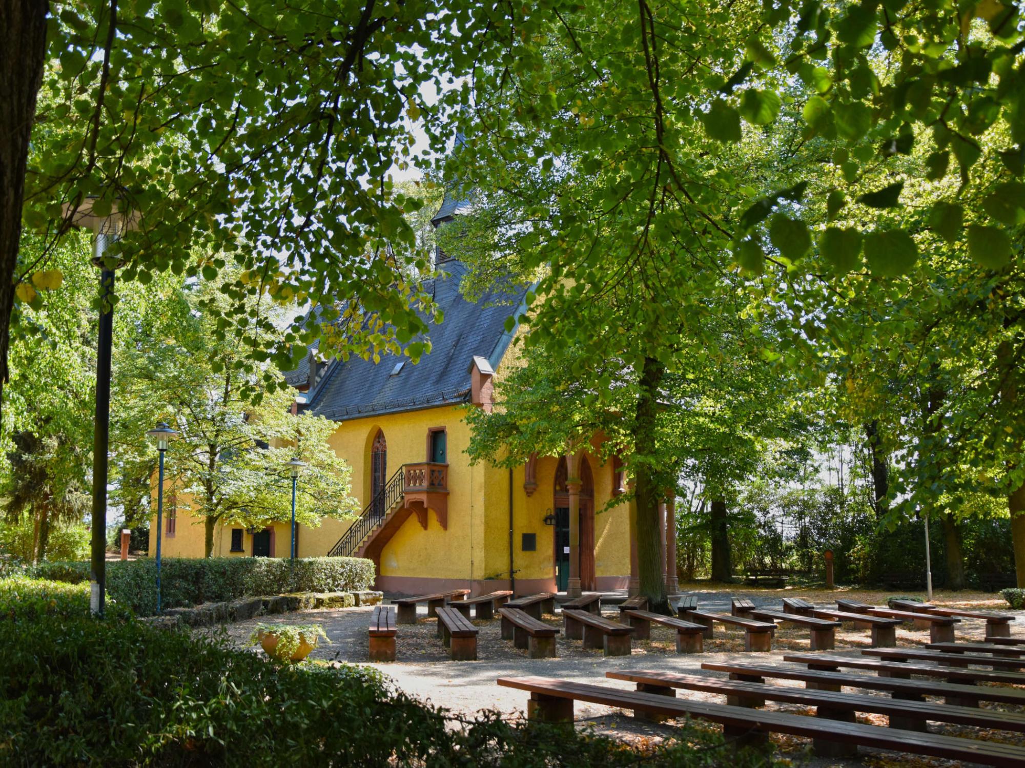 Die gelbe Kapelle versteckt hinter Laubbäumen im Sommer mit vielen Holzbänken davor harmonisch angeordnet