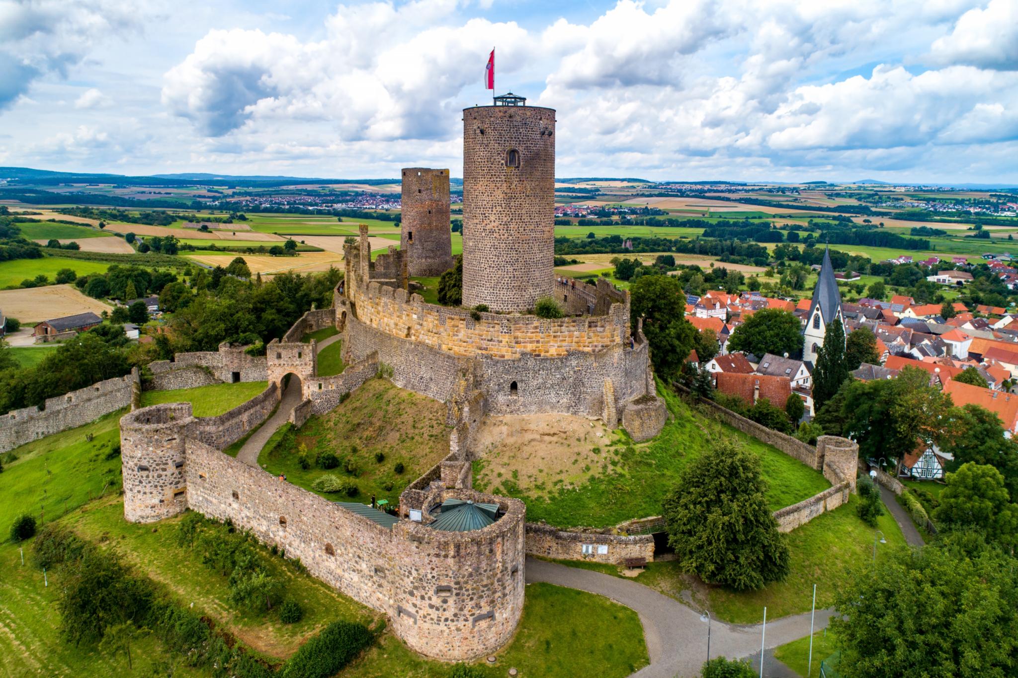 Das Bild zeigt die Burgruine Münzenberg mit den verschiedenen Wehrmauern und einem runden Turm. Drumherum sind Felder und Wiesen, auf der linken Seite ist die Ortschaft Münzenberg mit einigen Häusern zu erkennen.