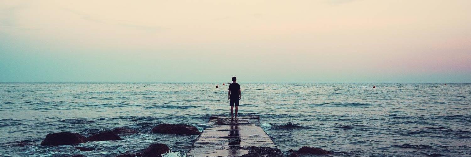 Person steht am Ende einer Buhne mit Blick auf das Meer und dem Horizont. Die Füße werden schon vom Meer umspielt. Im Meer schwimmen in der Ferne weitere Personen.