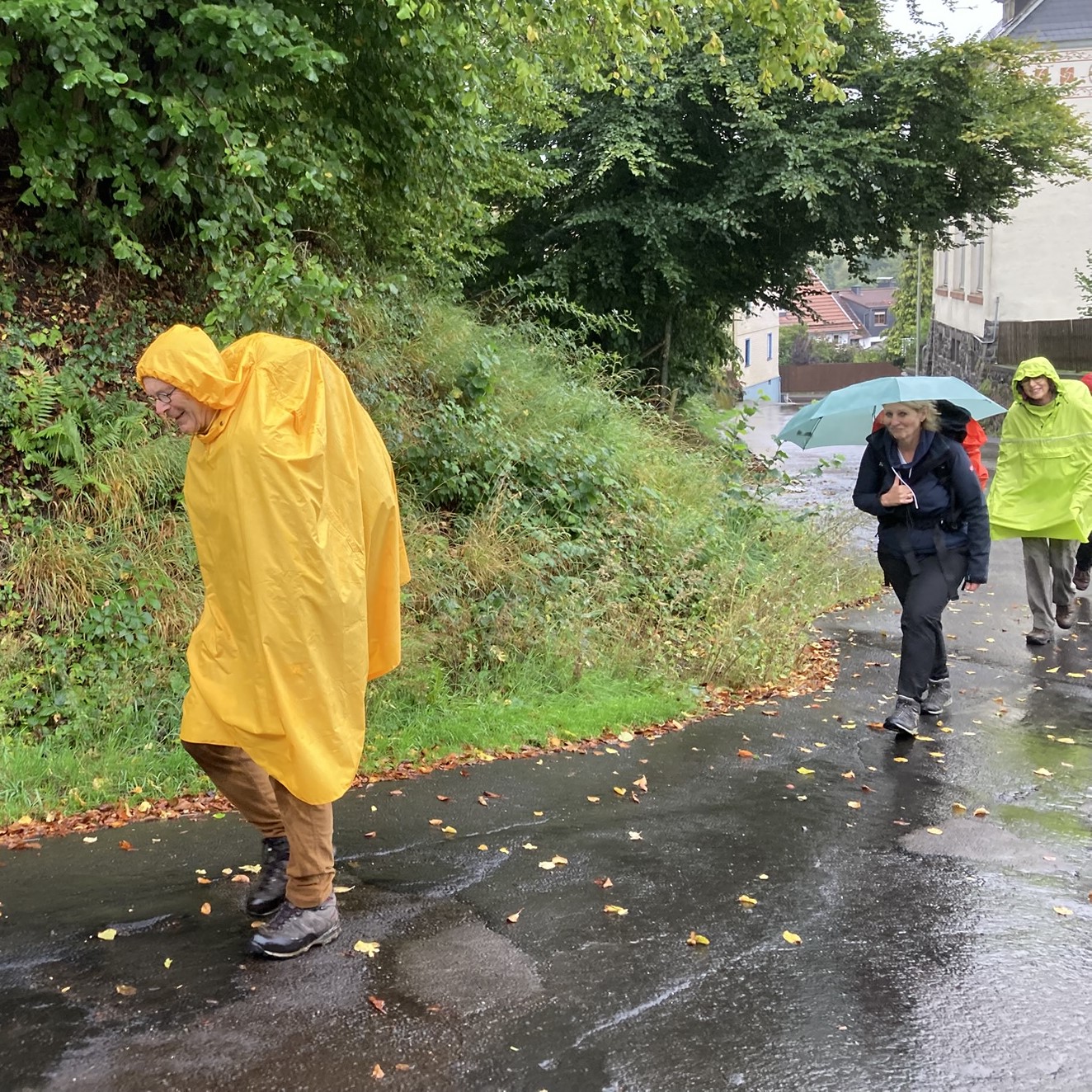 Wandergruppe mit Mann im gelben Regenponcho auf nassem Weg, eine Frau mit Schirm und Rucksack, eine weitere Frau mit grünem Poncho