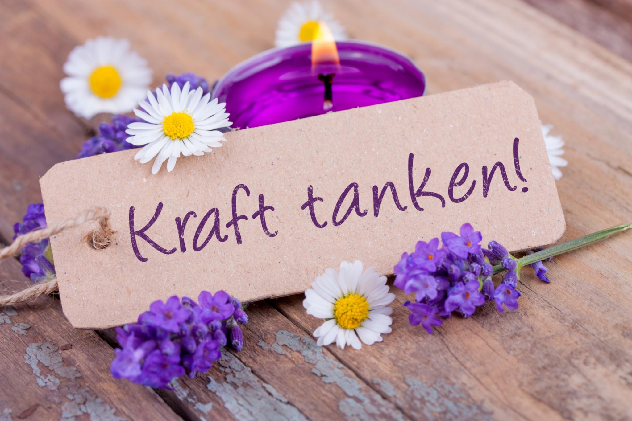 Ein kleines Anhängerschild mit den Worten „Kraft tanken“. Das Schild liegt gegen ein lila Teelicht gelehnt von Gänseblümchen und weiteren Blumen umrankt auf einem alten Holztisch.