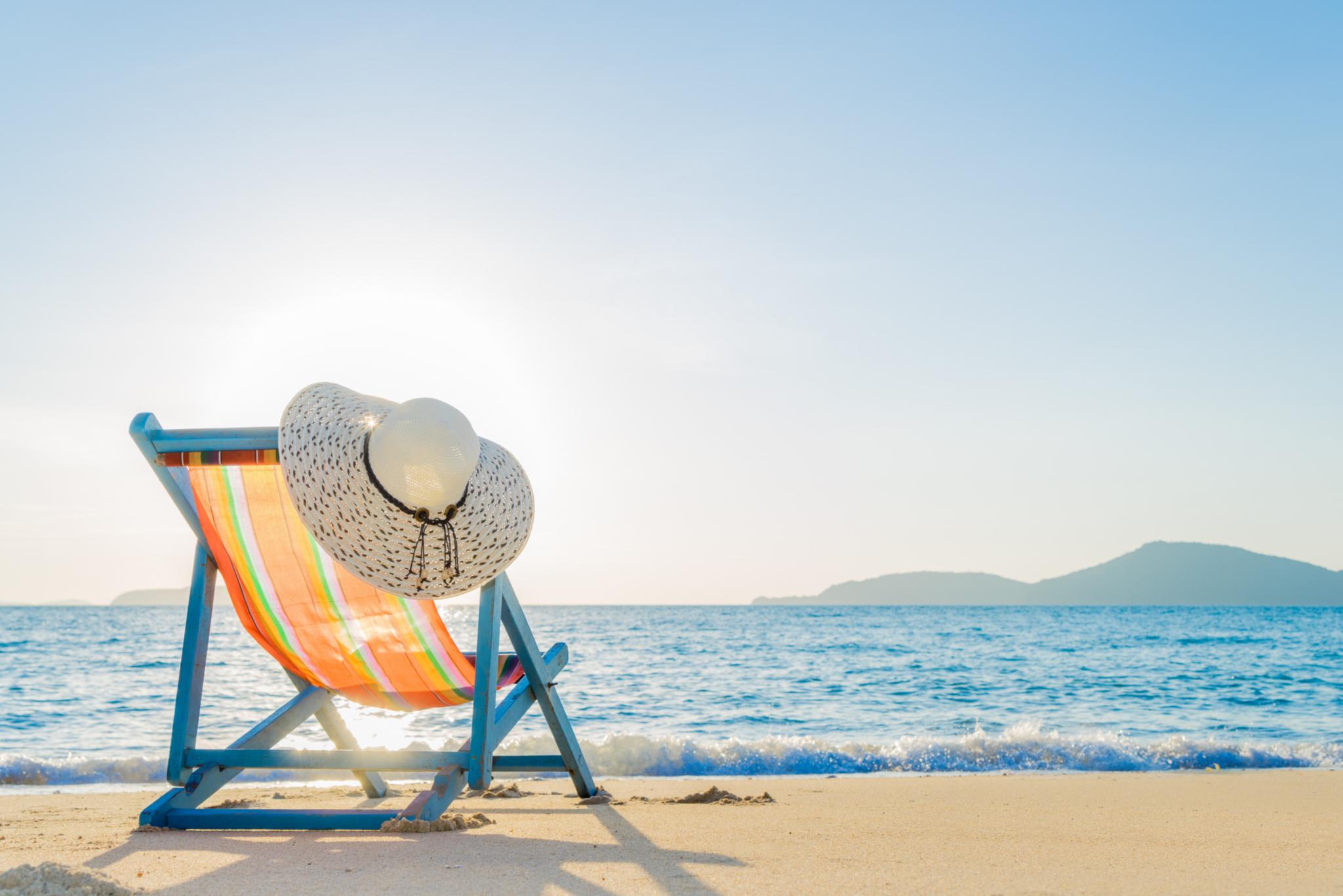 Ein Liegestuhl auf dem Strand stehend, das weite Meer, blauer Himmel, im Hintergrund eine Küste mit Bergen. An der Lehne des Stuhls hängt ein Sonnenhut.