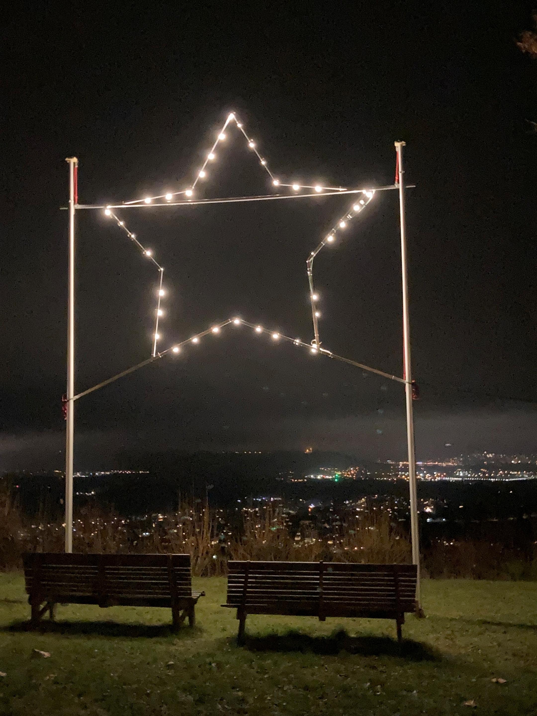 Stern aus Glühbirnen bei Nacht, im Hintergrund die Lichter von Gau-Algesheim und dem Rheingau, im Vordergrund zwei leere Bänke