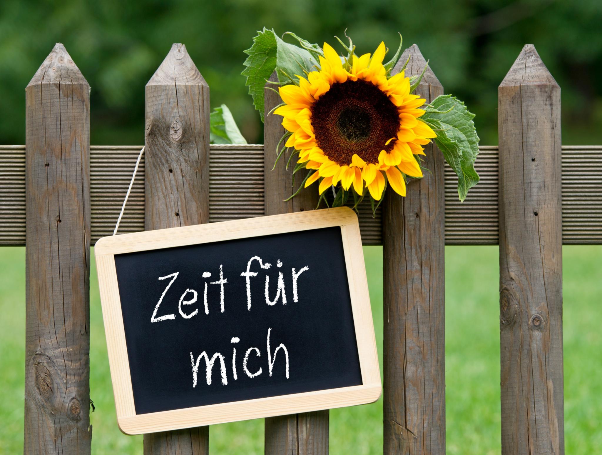 An einem Jägerzaun hängt eine kleine Kreidetafel mit der Aufschrift „Zeit für mich“. Eine Sonnenblume schaut darüber über den Zaun.