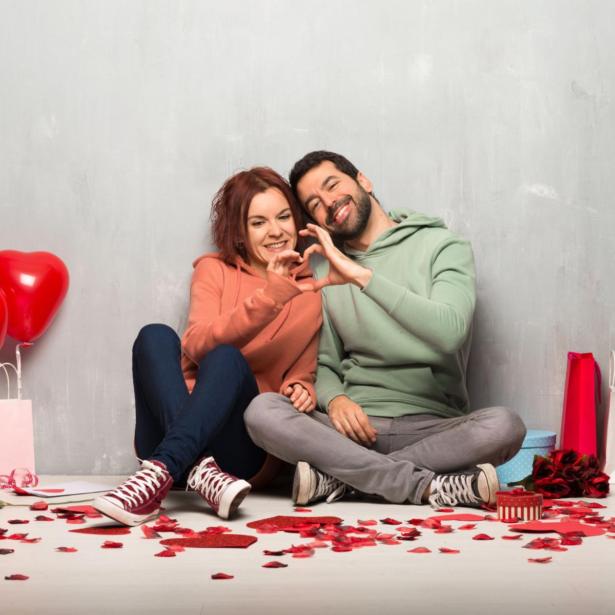 Paar am Valentinstag nebeneinander auf dem Boden sitzend machen ein Herz mit ihren Händen