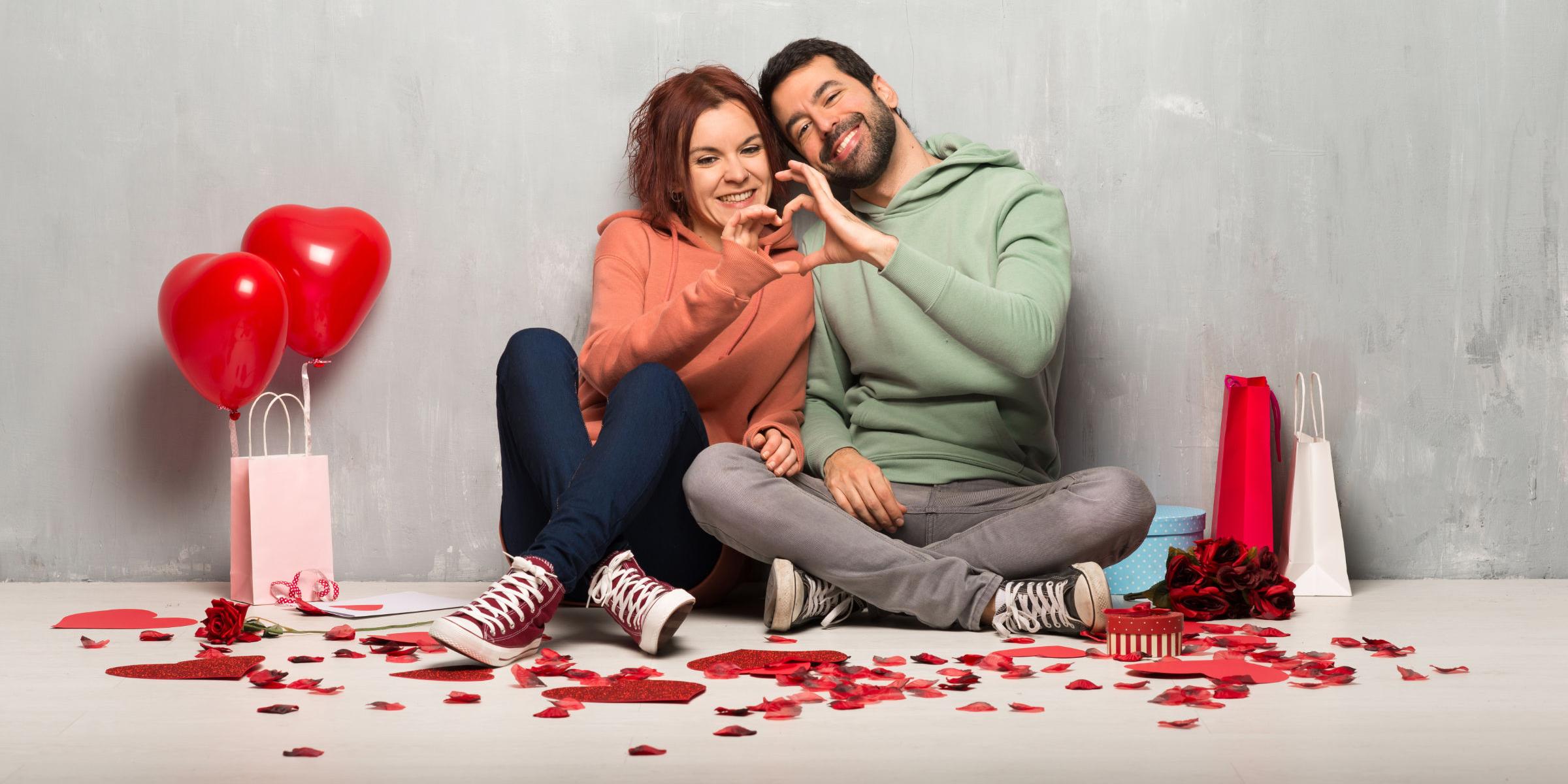 Paar am Valentinstag nebeneinander auf dem Boden sitzend machen ein Herz mit ihren Händen