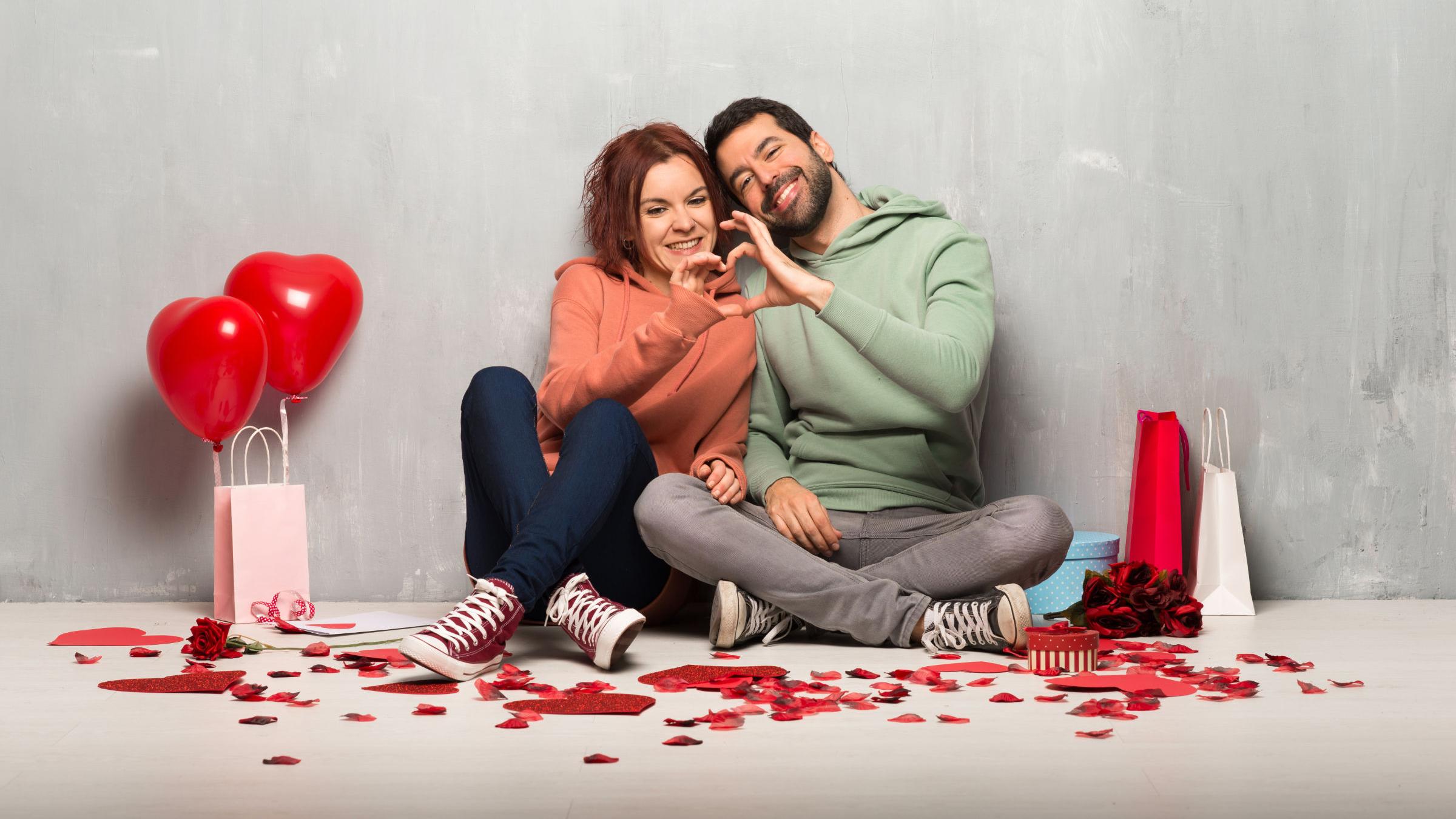 Paar am Valentinstag nebeneinander auf dem Boden sitzend machen ein Herz mit ihren Händen