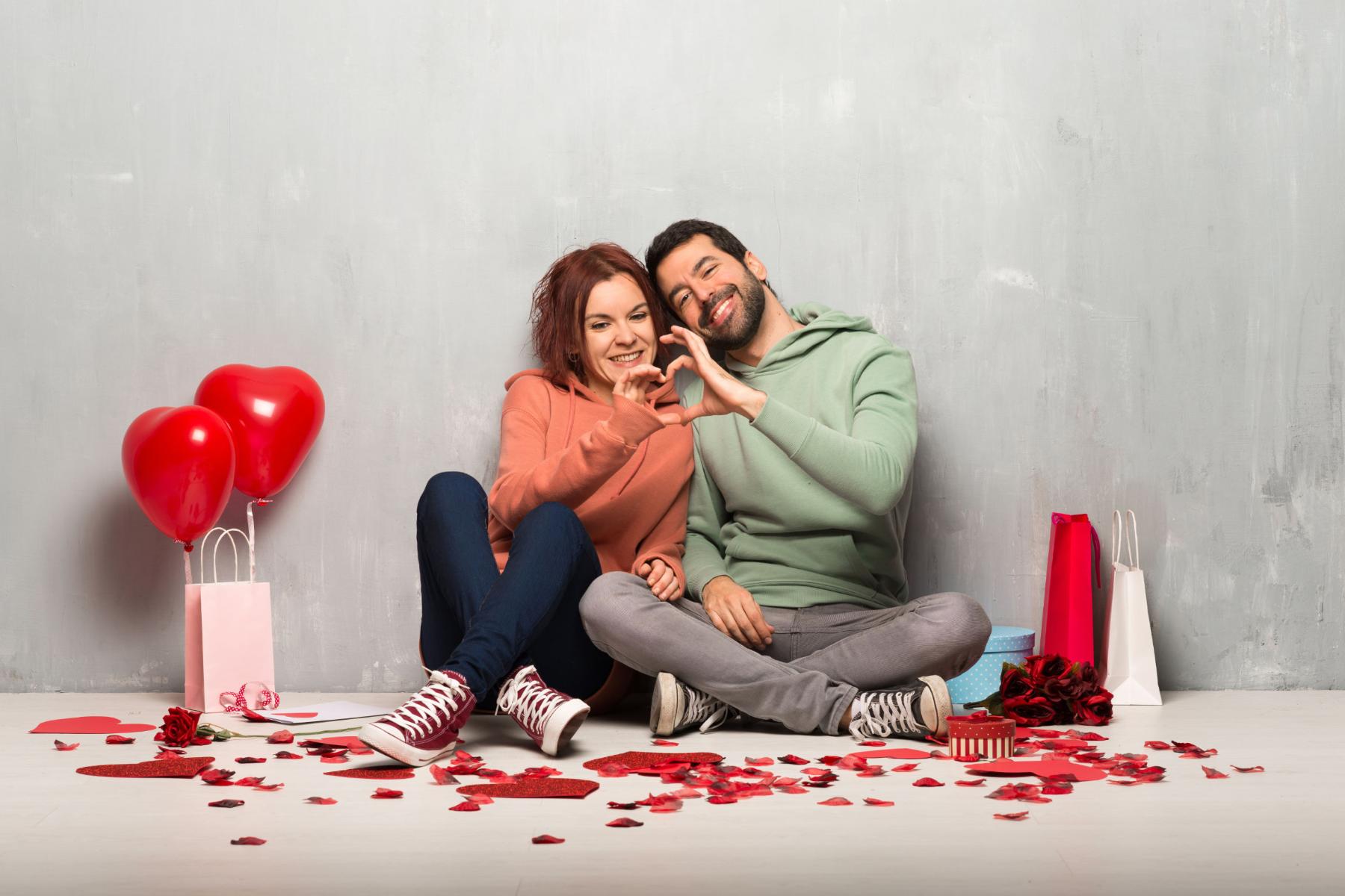 Paar am Valentinstag nebeneinander auf dem Boden sitzend machen ein Herz mit ihren Händen