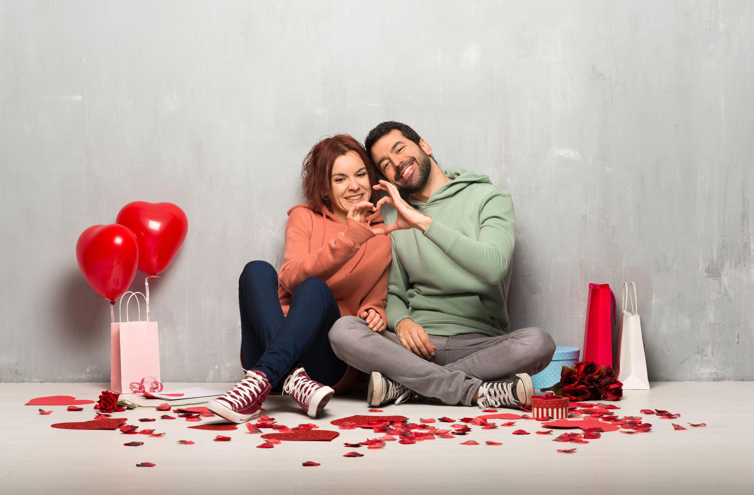 Paar am Valentinstag nebeneinander auf dem Boden sitzend machen ein Herz mit ihren Händen