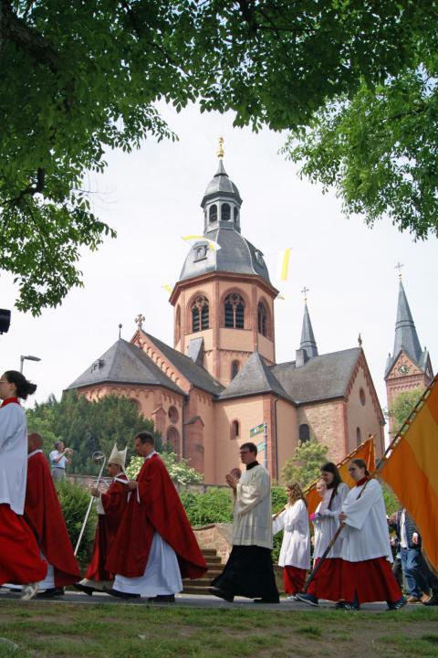 Auf dem Bild sieht man einen Einzug von Ministrant:innen und Priestern. Im Hintergrund ist die Seligenstädter Basilika, gerahmt von Bäumen zu erkennen.