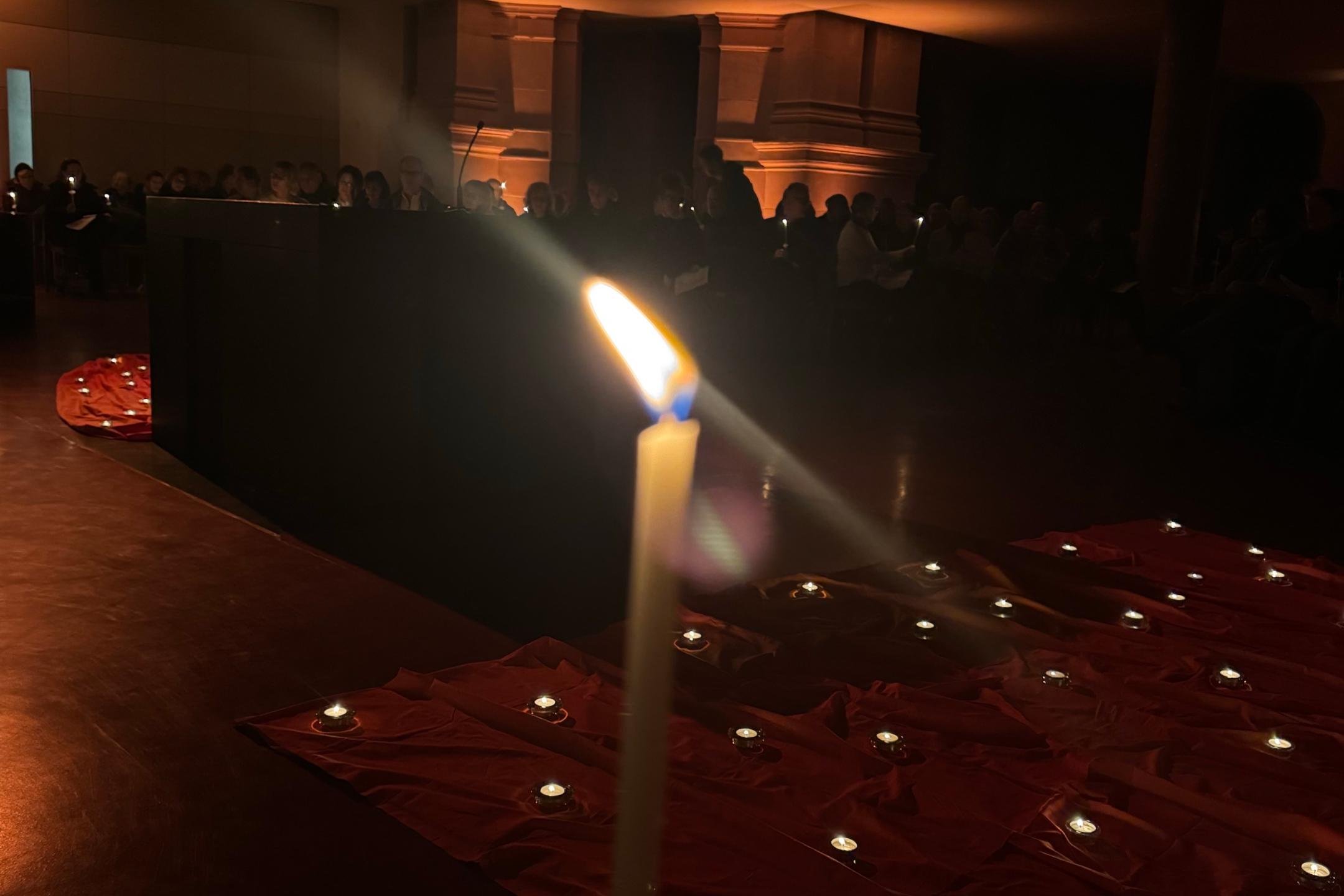 Eine Taizékerze brennt im Vordergrund. Dahinter sieht man die orangenen Tücher mit Teelichtern und im Hintergrund einen Teil der Gottesdienstbesucher:innen sowie die Beleuchtung der Kirche.