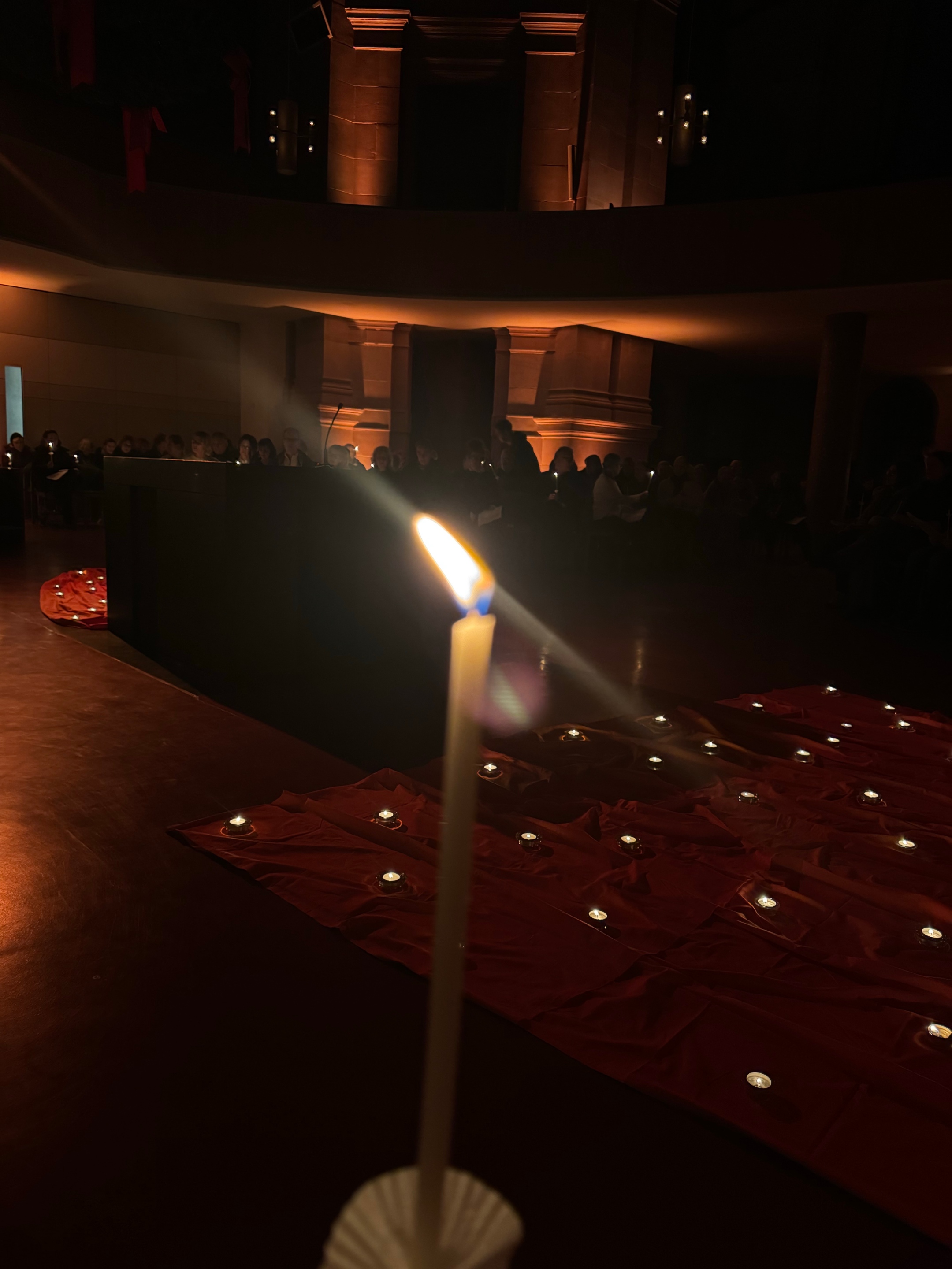 Eine Taizékerze brennt im Vordergrund. Dahinter sieht man die orangenen Tücher mit Teelichtern und im Hintergrund einen Teil der Gottesdienstbesucher:innen sowie die Beleuchtung der Kirche.