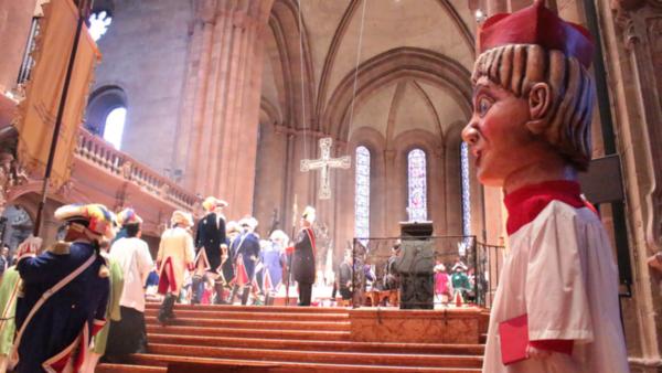 Der Mainzer Dom beim Gardegottesdienst, Menschen gehen in Fastnachtskostümen hoch zum Altarraum, rechts steht ein Mensch in liturgischem Gewand und mit einer Schwellkopf-Verkleidung