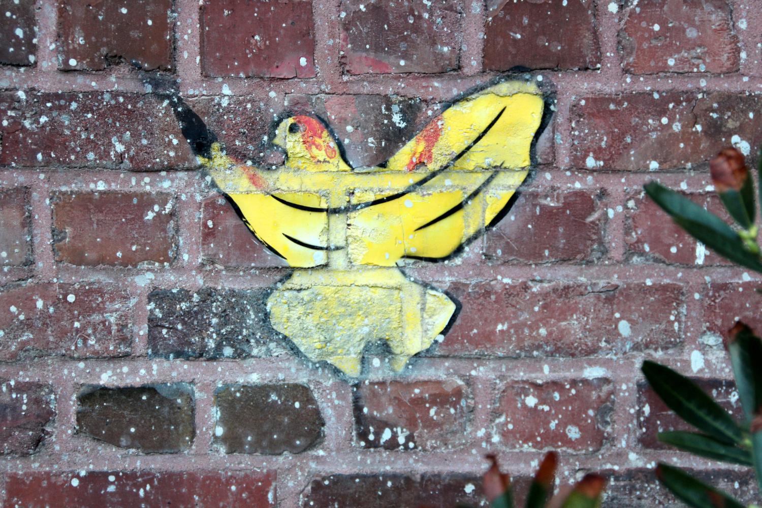 Gelbes Schmetterlings-Graffiti auf roter Backsteinmauer, daneben grüne Blätter