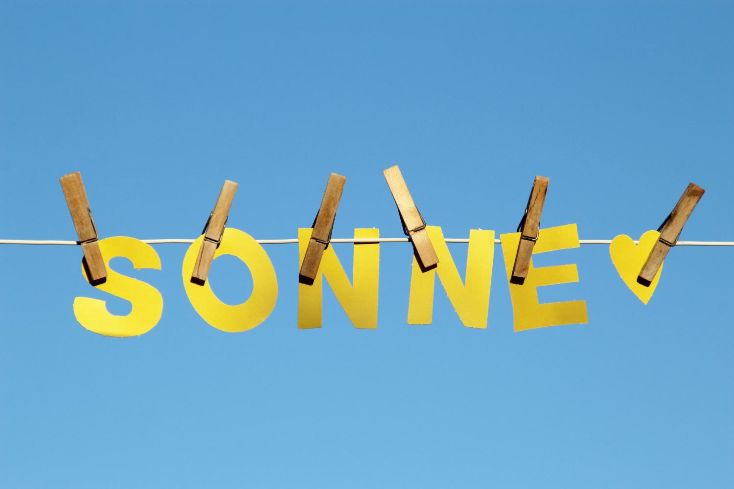 Aus gelbem Papier ausgeschnittenen Buchstaben des Wortes 'SONNE' und ein kleines gelbes Herz hängen mit hölzernen Wäscheklammern an einer weißen Wäscheleine vor einem blauen, wolkenlosen Himmel