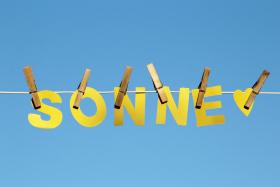 Aus gelbem Papier ausgeschnittenen Buchstaben des Wortes 'SONNE' und ein kleines gelbes Herz hängen mit hölzernen Wäscheklammern an einer weißen Wäscheleine vor einem blauen, wolkenlosen Himmel