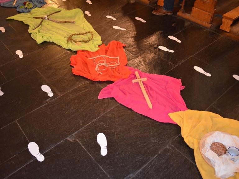 Bunte Tücher, ausgebreitet auf Kirchenfußboden, darauf Gegenstände als Symbole für die Kreuzwegstationen Jesu. Daneben liegen aus Papier ausgeschnittene Fußspuren.