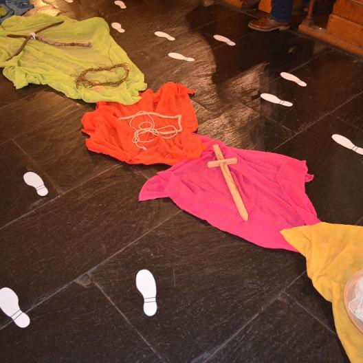 Bunte Tücher, ausgebreitet auf Kirchenfußboden, darauf Gegenstände als Symbole für die Kreuzwegstationen Jesu. Daneben liegen aus Papier ausgeschnittene Fußspuren.