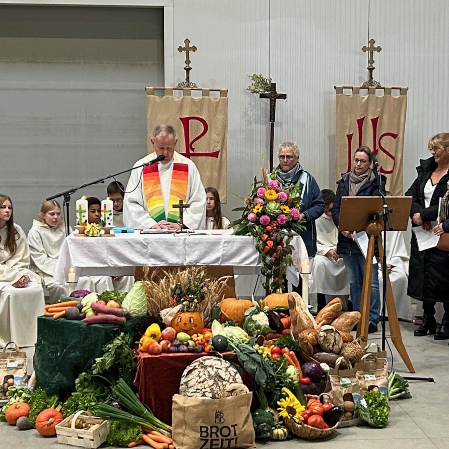 Einfacher Altar in der 'Kartoffelhalle' vor dem Altar viele Erntegaben, Obst, Gemüse, Brot, dahinter Pfr Swiatek mit Regenbogenstola