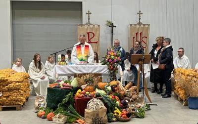 Einfacher Altar in der 'Kartoffelhalle' vor dem Altar viele Erntegaben, Obst, Gemüse, Brot, dahinter Pfr Swiatek mit Regenbogenstola