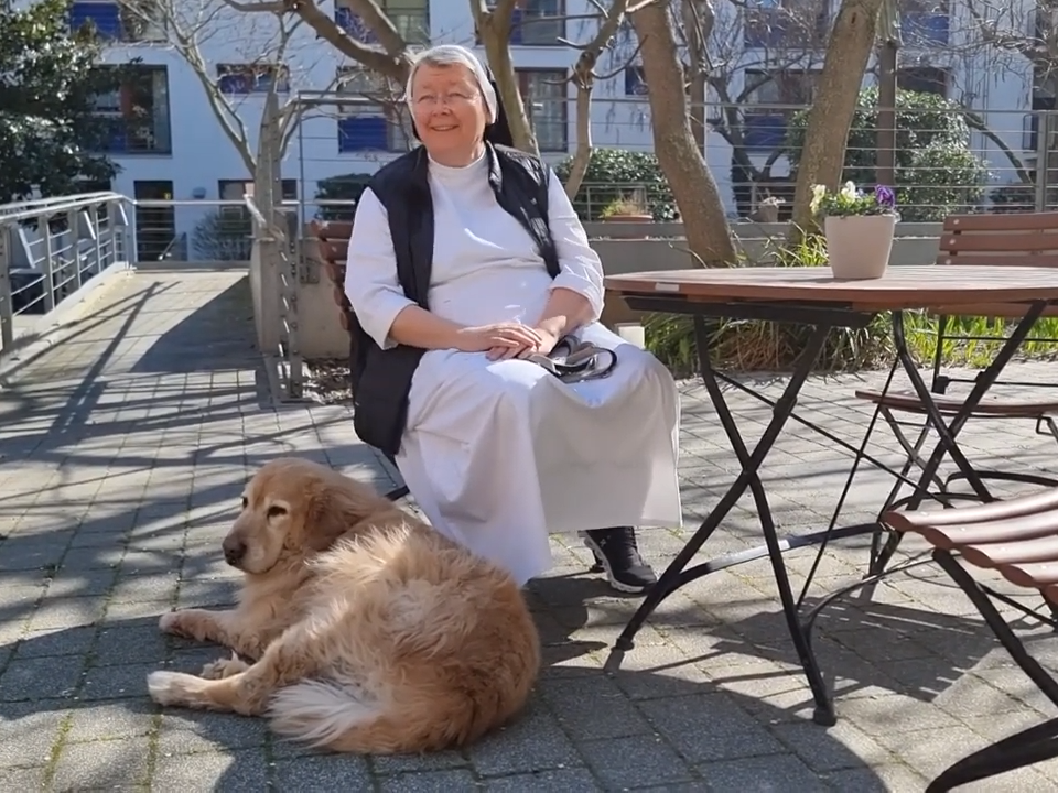 Ordensschwester am Gartentisch mit Hund Henri, der entspannt zu ihren Füßen liegt
