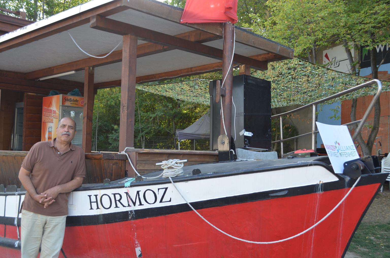 Behrouz Asadi vor dem ausrangierten Rheinschiff, das im Freigelände des „Hauses der Kulturen“ für den Getränkeausschank genutzt wird. Es ist nach seinem verstorbenen Bruder Hormoz benannt. (c) Maria Weissenberger