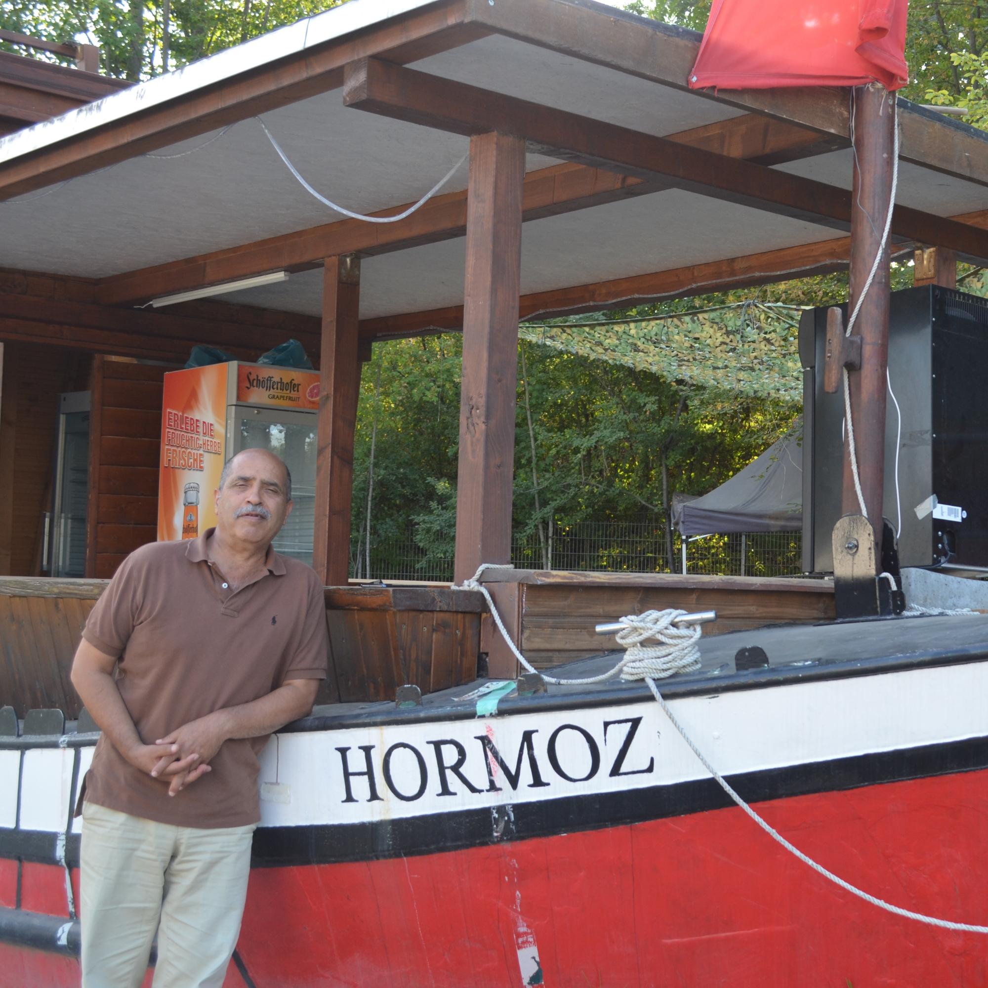 Behrouz Asadi vor dem ausrangierten Rheinschiff, das im Freigelände des „Hauses der Kulturen“ für den Getränkeausschank genutzt wird. Es ist nach seinem verstorbenen Bruder Hormoz benannt.