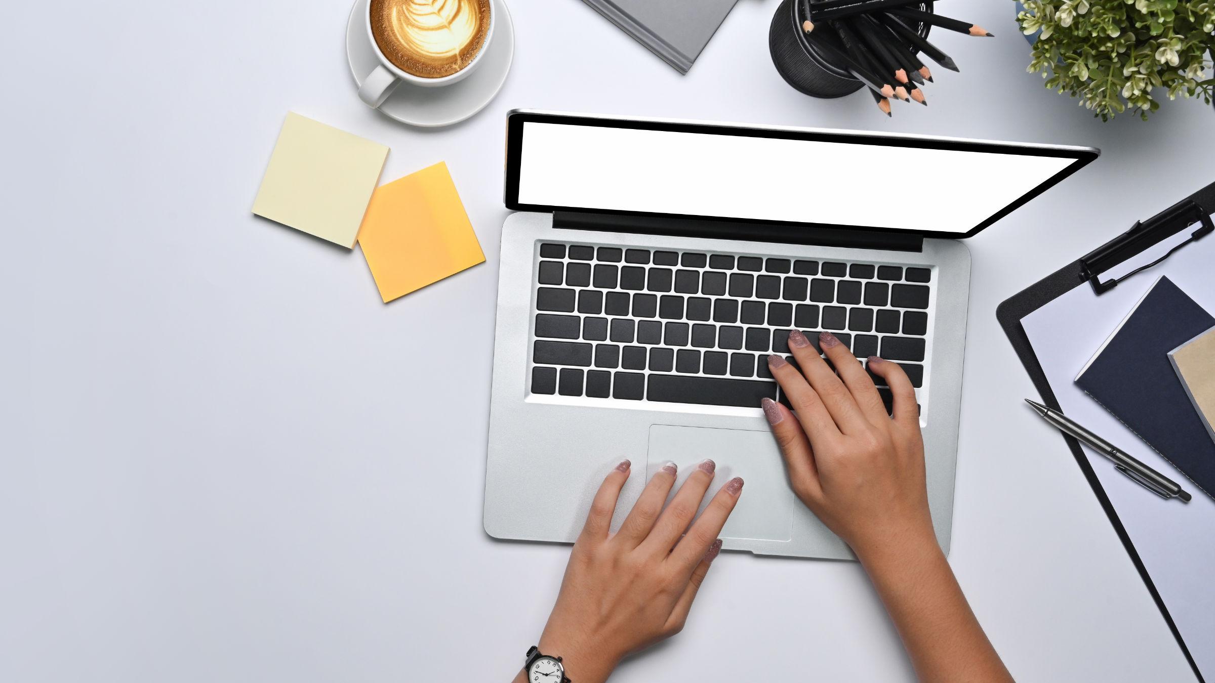 Blick von oben auf einen Laptop und die Arme einer Frau, die daran arbeitet. Der Laptop steht auf einem weißen Tisch mit Capucchino, Pflanze, Bleistiften und einer Armbanduhr.