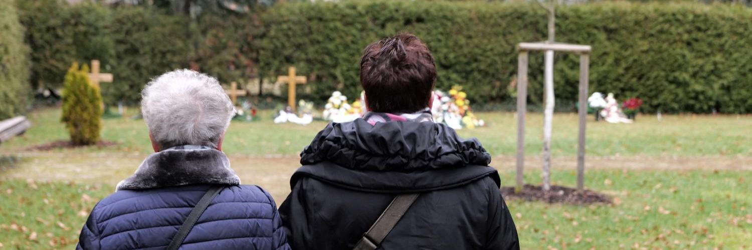 Zwei Frauen stehen trauernd vor frischen Gräber mit Holzkreuzen.