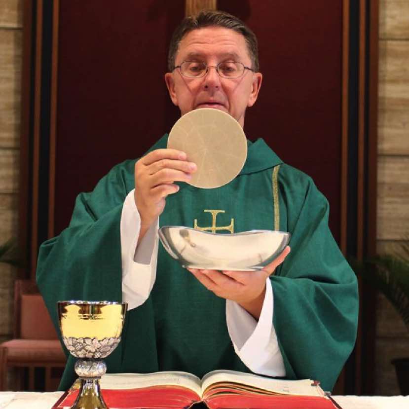 Pfarrer in grünem Messgewand am Altar, hält große Hostie und Hostienschale hoch.