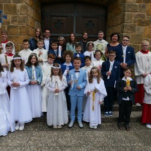 Gruppe von 17 Kindern in weißen Kleidern und festlichen Anzügen mit großen Kerzen, Messdiener und einige Erwachsene vor einer Kirchentür.