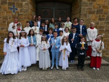 Gruppe von 17 Kindern in weißen Kleidern und festlichen Anzügen mit großen Kerzen, Messdiener und einige Erwachsene vor einer Kirchentür.