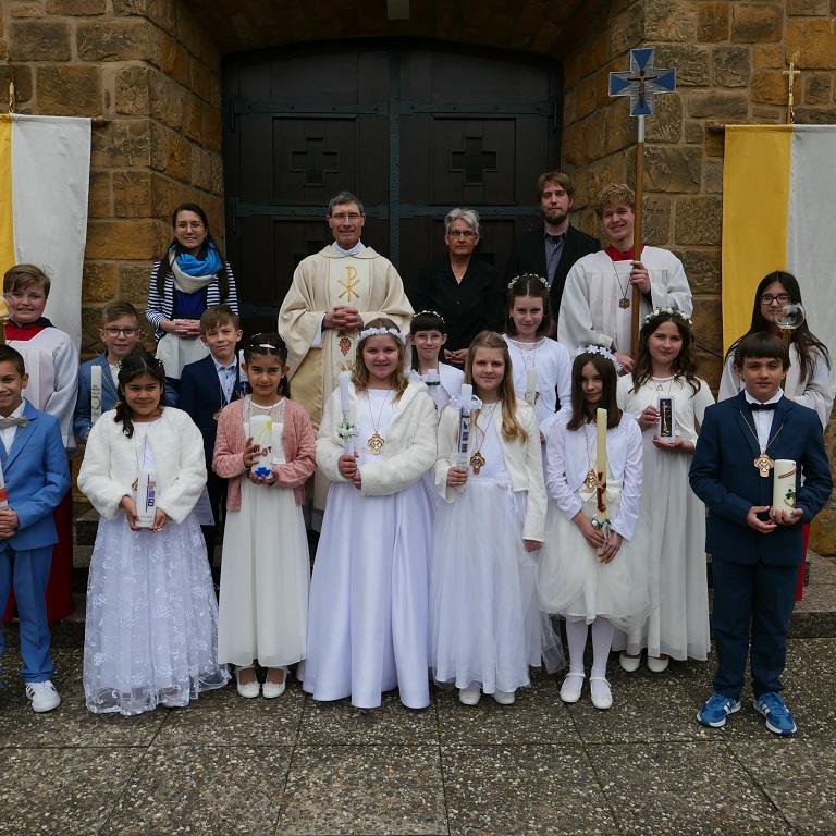 12 Erstkommunionkinder in weißen Kleidern und dunklen Anzügen mit Kerzen in den Händen mit Pfarrer, weiteren Erwachsenen und Messdienern vor einer Kirchentür.