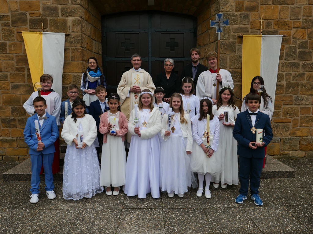 12 Erstkommunionkinder in weißen Kleidern und dunklen Anzügen mit Kerzen in den Händen mit Pfarrer, weiteren Erwachsenen und Messdienern vor einer Kirchentür.