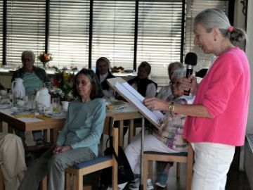 Eine Frau am Rednerpult spricht über ein Mikro zu Frauen, die an Tischen sitzen.