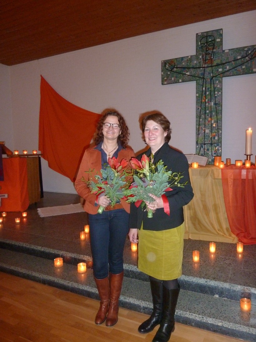 Zwei Frauen mit Blumen in der Hand stehen vor einem Altar und lächeln freundlich in die Kamera.