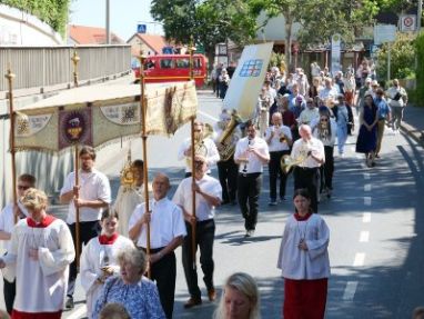 Viele Menschen in einer Prozession auf einer Straße mit einem Baldachin und darunter ein Priester mit einer Monstranz.