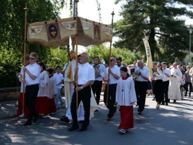 Priester mit Monstranz unter einem Baldachin, der von vier Männern getragen wird und weitere Menschen.