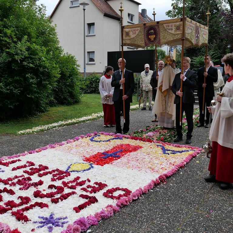 Ein Priester mit einem goldenen Gefäß unter einem Baldachin, der von vier Männern getragen wird, vor einem großen Blumenteppich.
