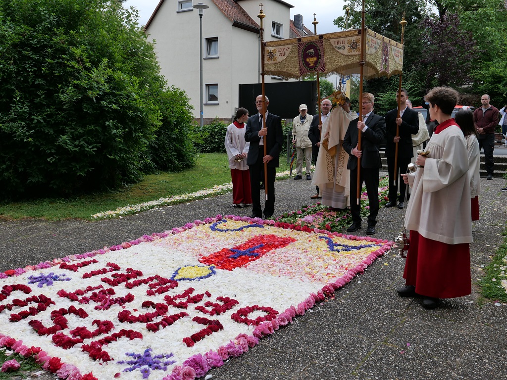 Ein Priester mit einem goldenen Gefäß unter einem Baldachin, der von vier Männern getragen wird, vor einem großen Blumenteppich.