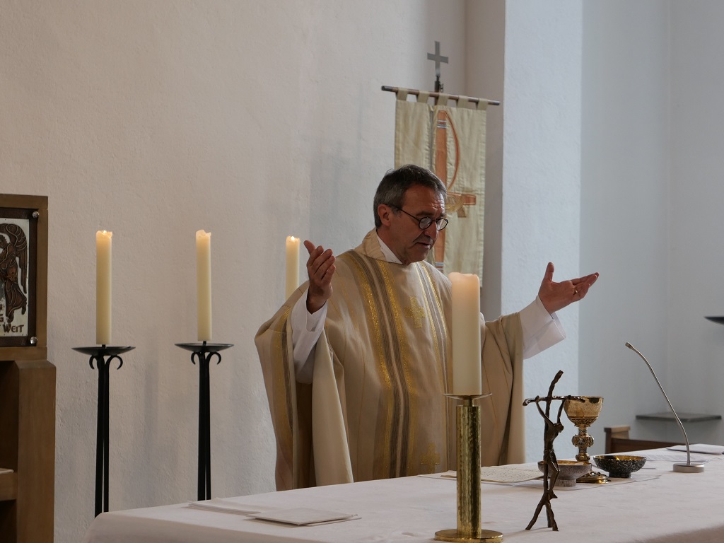 Ein Priester in weißem Messgewand an einem Altar mit ausgebreiteten Händen.