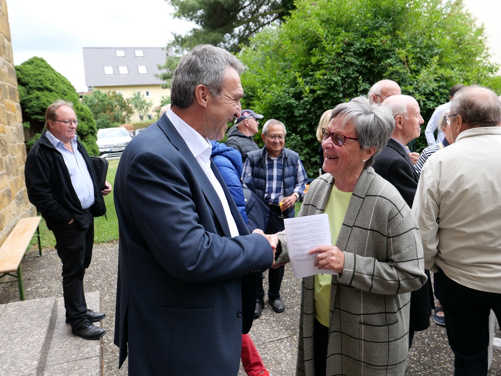 Eine Frau schüttelt einem Mann die hand, weitere Menschen stehen im Hintergrund.