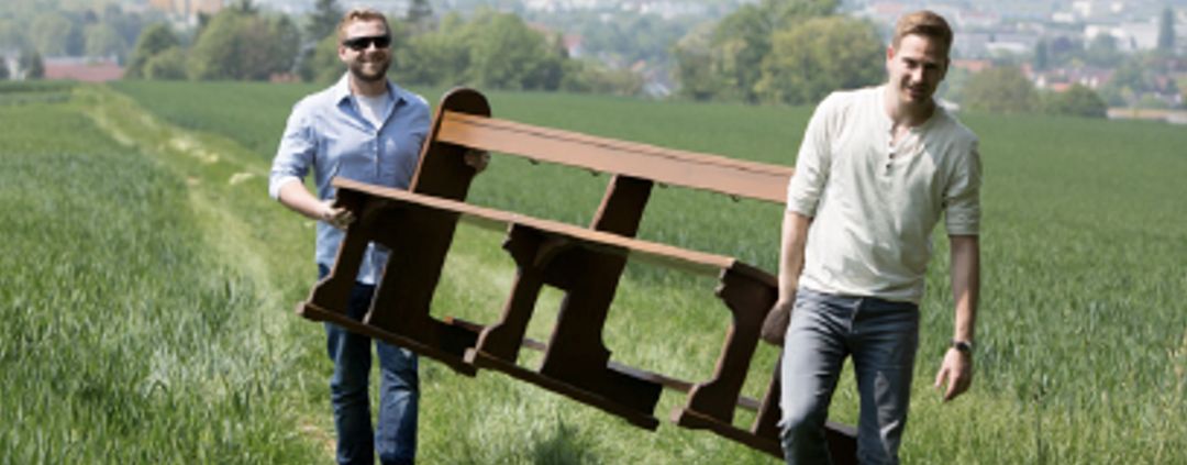 Zwei Männer tragen eine Kirchenbank über einen Feldweg.