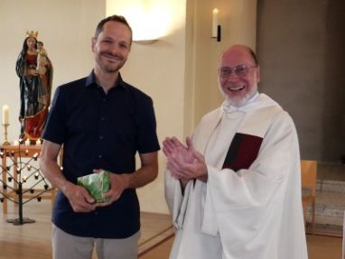 Ein Priester im Messgewand und ein Mann mit einem Geschenk in der Hand stehen in einem Kirchenraum.