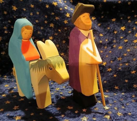 Abgebildet sind zwei hölzerne Krippengfiguren: Maria auf dem Esel und Josef mit einem Stab in der Hand